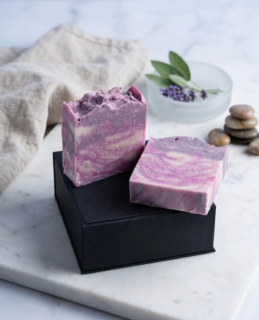 Two purple swirled soap bars on a black stand with a marble surface and lavender flowers in the background.