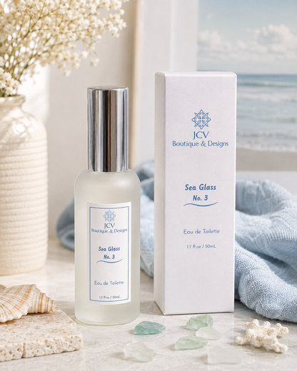 A bottle of sea glass no. 3 eau de toilette by JCV Boutique & Designs next to its white packaging box, styled with seashells, sea glass, and a blue towel on a light stone surface.