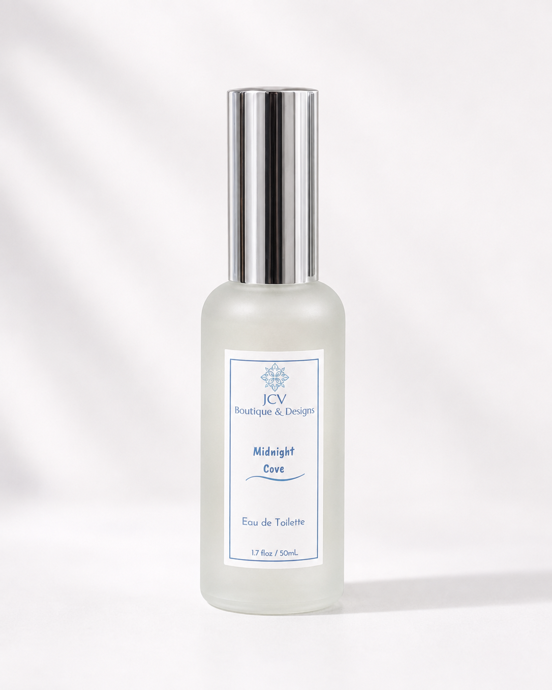 A frosted glass perfume bottle with a shiny silver cap, featuring a white label with blue text that reads 'JCV Boutique & Design Midnight Cavi Eau de Toilette'. The bottle is centered against a clean white background with soft, artistic leaf shadows.