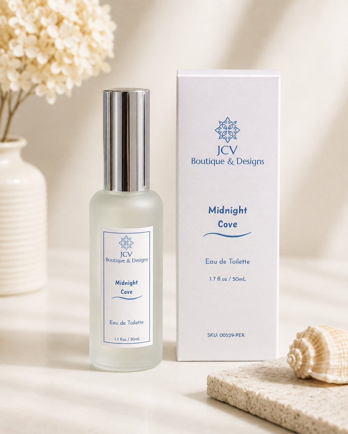 A bottle of JCV Boutique & Designs Midnight Cove Eau de Toilette next to its white packaging box, styled on a neutral beige surface with dried flowers and a seashell in the background.