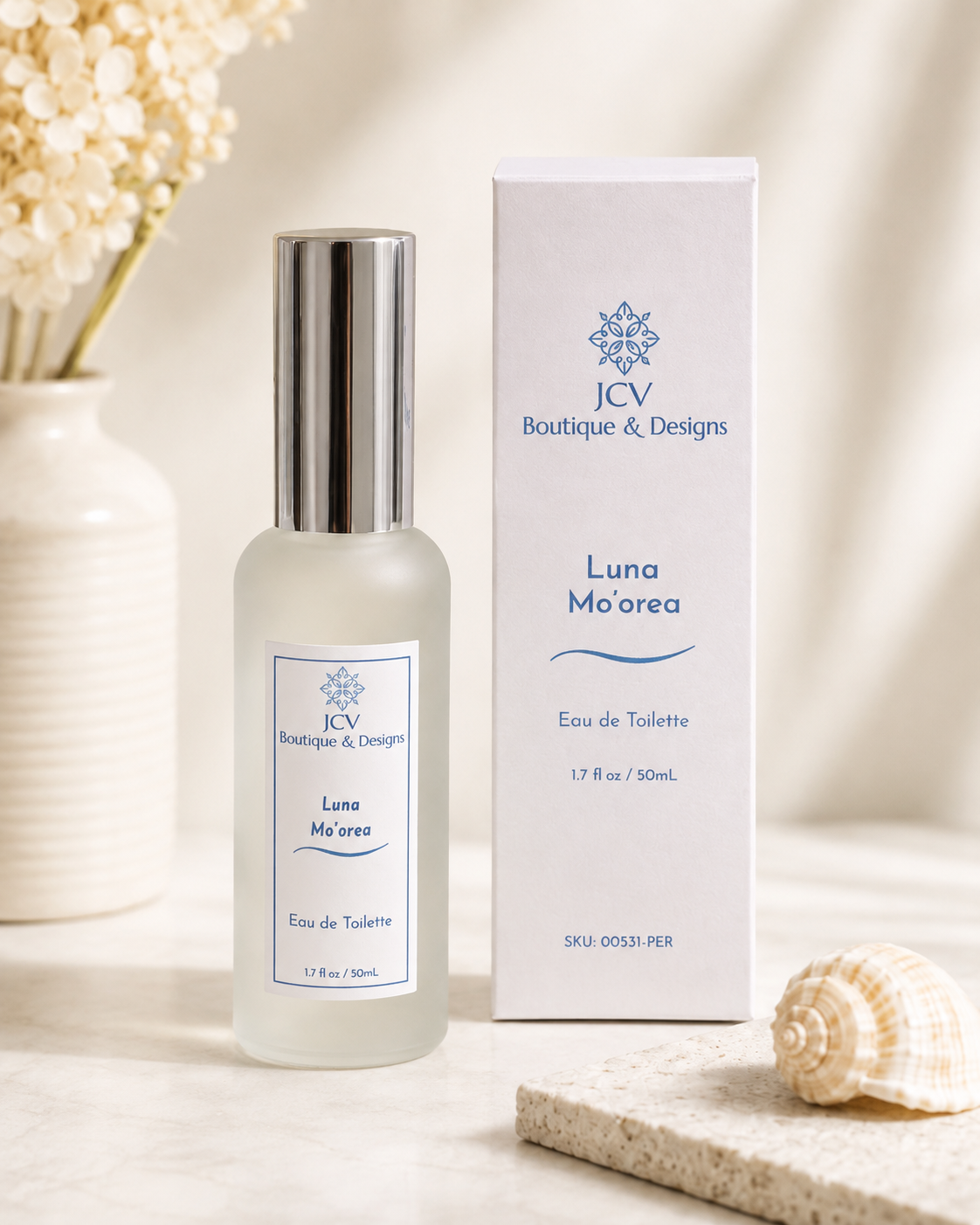 A bottle of JCV Boutique & Designs Luna Mo'orea Eau de Toilette with a silver cap stands next to its white packaging box on a light marble surface, styled with a seashell and dried flowers.