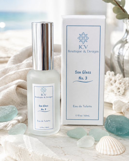 A frosted glass perfume bottle with a silver cap stands beside its white box, both labeled 'ICV Boutique & Designs Sea Glass No. 3 Eau de Toilette,' styled on a marble surface with sea glass and a seashell against a soft beach background.