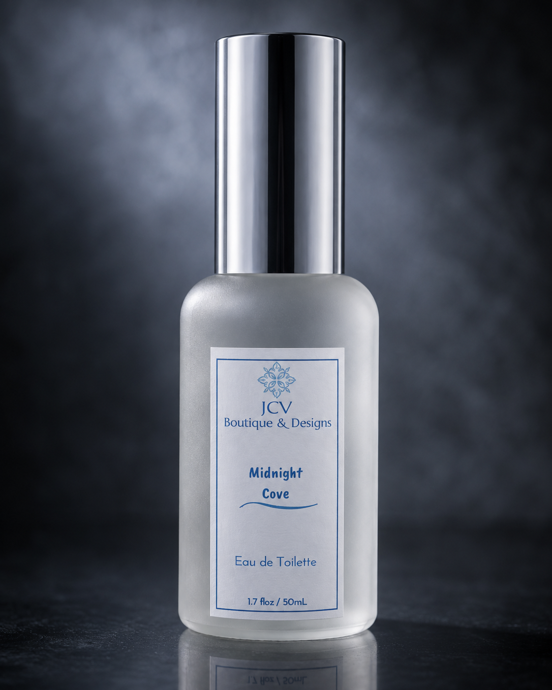 A frosted glass perfume bottle with a shiny silver cap, featuring a white label with blue text that reads 'JCV Boutique & Designs Midnight Cove Eau de Toilette'. The bottle is centered against a dark, smoky gray background with a soft reflection below.