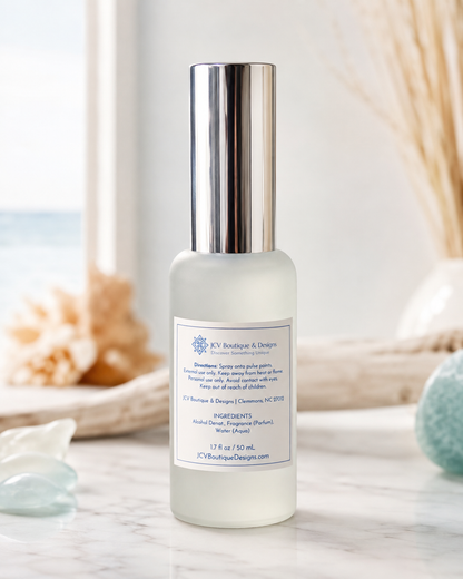 A frosted glass bottle of body oil with a metallic silver cap, standing on a marble surface with a blurred beach and dried grass background.