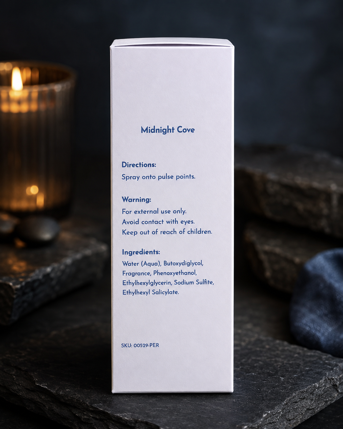 A tall white rectangular product box standing upright on a dark stone surface, with printed blue text reading 'Midnight Cove' and usage directions. A blurred lit candle and dark background are visible behind it.