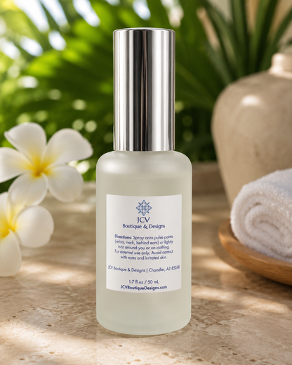 A frosted glass bottle of body wash with a reflective silver cap, labeled 'JCW Boutique & Designs', set on a stone surface with white flowers and a rolled towel in the background.