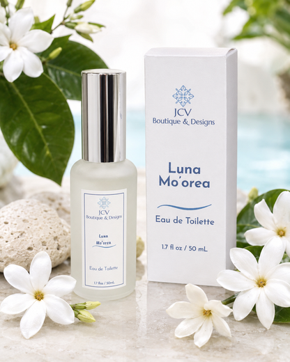 A bottle of JCV Boutique & Designs Luna Mo'orea Eau de Toilette with a silver cap stands next to its white box, styled with white flowers and green leaves on a light stone surface.