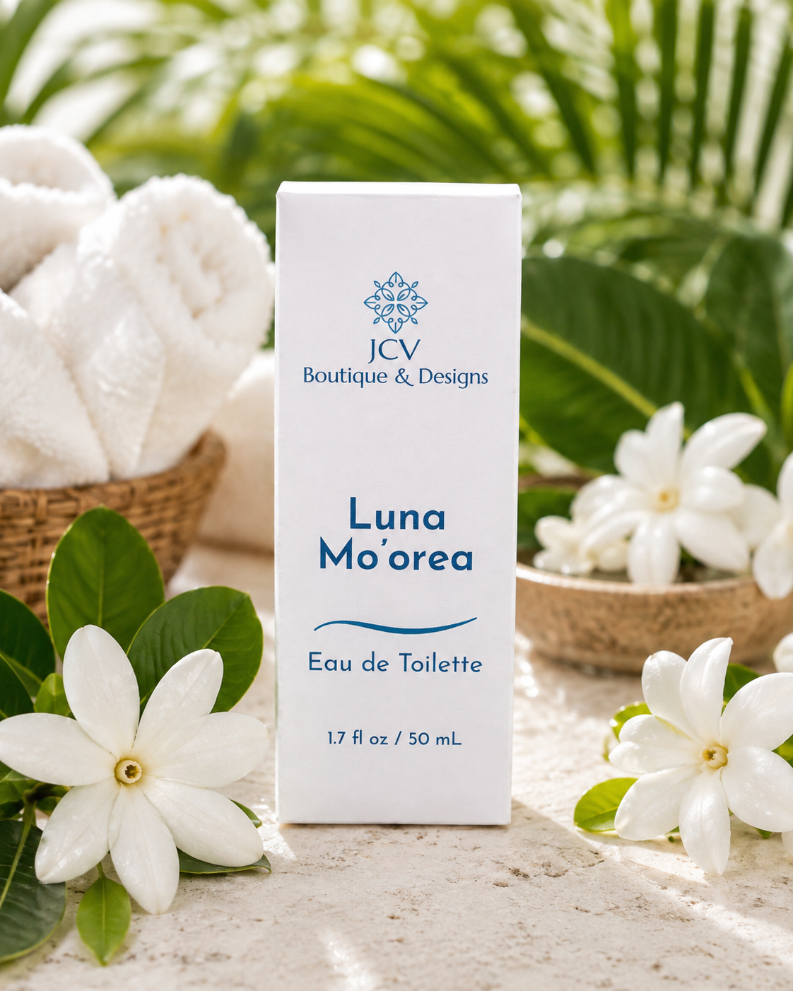 A white rectangular box labeled 'JCV Boutique & Designs Luna Mo'orea Eau de Toilette 1.7 fl oz / 50 mL' stands on a light stone surface, surrounded by white flowers, green leaves, folded white towels, and tropical foliage in the background.